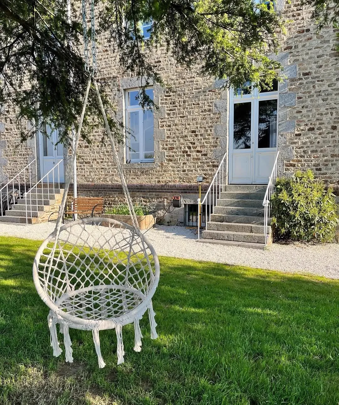 Fauteuil suspendu en macramé sur la pelouse devant la façade en pierre du Manoir Ducey